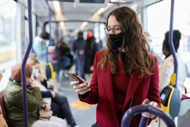 Femme masque noir manteau et pull rouge avec lunette dans bus bleu regardant son téléphone portable
